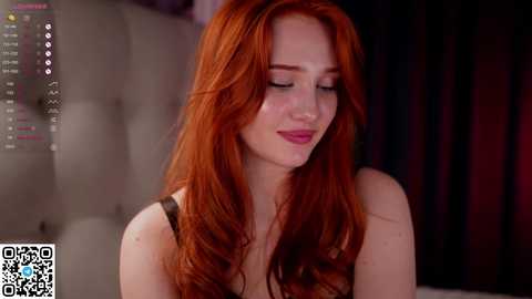 Media: Video of a fair-skinned, red-haired woman with long, wavy hair, wearing a black top, smiling softly. Background shows a blurred, dimly-lit room with a grey cushioned headboard.