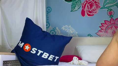 A video of a cozy bedroom featuring a navy blue pillow with \"MSTBET\" text and a red star, surrounded by white bedding and a light blue wall with floral patterns.