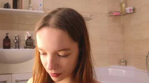 Video of a young Caucasian woman with long, straight brown hair, gazing downward in a beige-tiled bathroom. The background includes a white sink, bottles, and a bathtub.