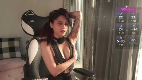 Video of a woman with medium skin tone, dark hair, and tattoos, wearing a black halter top, seated in a black and white gaming chair, with a fitness app displaying stats on a screen in the background.