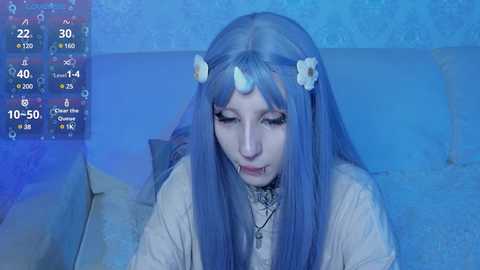 Video of a pale-skinned woman with long, straight blue hair, wearing a white floral crown, a white dress, and a black choker, seated on a blue couch in a dimly lit room.