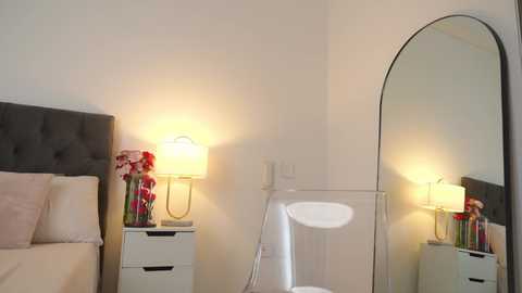 Media: Video of a minimalist, modern bedroom with a white toilet, beige bed, grey tufted headboard, white nightstand with lamp and red flowers, large round mirror, and off-white walls.