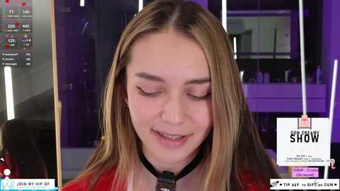 Media: Video of a young woman with light skin and long blonde hair, wearing a black choker, smiling slightly. Background features a modern kitchen with purple lighting, and a \"SLOW\" sign.