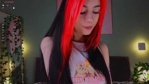 Video of a young woman with long, dyed red and black hair, wearing a sleeveless graphic tee, standing indoors in a softly lit room with green walls and leafy decorations.