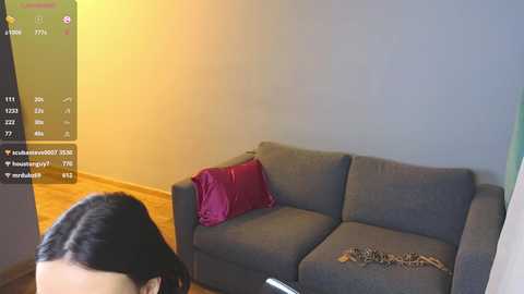 A video of a dark-haired woman in a gray sweater, sitting on a gray sofa with a red pillow, looking at a phone in a room with wooden flooring and beige walls.