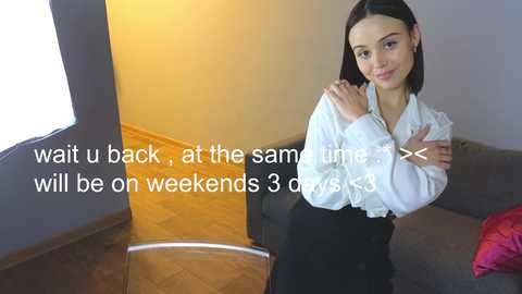 Media: Video of a young Asian woman with straight black hair, wearing a white blouse and black skirt, standing in a modern living room with wooden flooring, dark walls, and a grey couch. Text overlay reads: \"wait u back, at the same time will be on weekends 3 days\