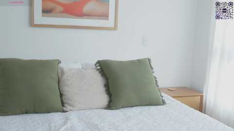 Media: Video of a minimalist bedroom with a white bed, two green and one beige pillow, a wooden nightstand, and a framed abstract painting on the white wall.