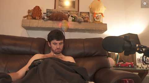 Media: Video of a bearded man in a brown leather couch, covered with a blanket, in a cozy living room with a fireplace, stuffed toys, and a lamp.