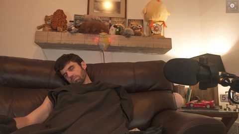 Media: Video of a man in a dark shirt lounging on a brown leather couch, with a plush toy dog on a fireplace mantle and a lamp on a wooden side table.