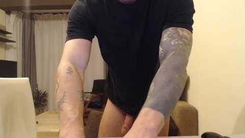 Media: Video of a muscular man with a black shirt and tattoos on his arms, standing in a dimly lit living room with beige walls, a TV, and a couch.