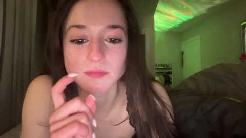 Media: Video of a young woman with fair skin and long brown hair, wearing a black top, gazing intently at her nails. Background features a dimly lit bedroom with green LED lighting and messy bedding.