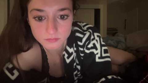 Media: A video of a young woman with long brown hair, wearing a black and white geometric-patterned robe, lying on a bed in a dimly lit bedroom.