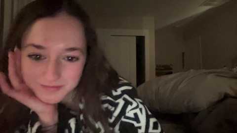 Media: A video of a young woman with curly brown hair, wearing a black and white patterned shirt, leaning on her hand, in a dimly lit bedroom with messy bedding and a door in the background.