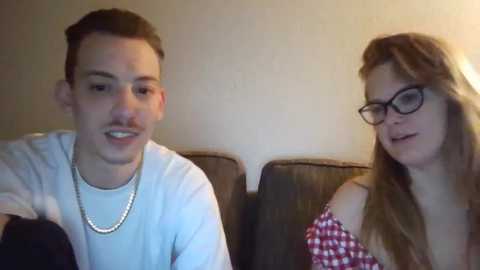 Media: Video of a young Caucasian couple sitting on a couch. Man with short brown hair, wearing a white t-shirt and silver chain necklace. Woman with long brown hair, glasses, and red checkered top. Plain beige wall background.