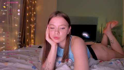 A video of a young, fair-skinned woman with long brown hair, lying on her stomach on a bed covered with a white, polka-dotted blanket. She wears a light blue tank top and black panties, with her legs bent and feet up. The room has a cozy, dimly lit atmosphere with fairy lights and a TV in the background.