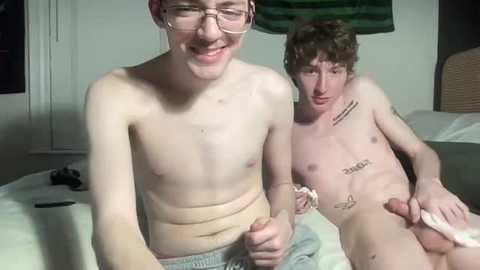 Media: Video of two young, slender, naked men with pale skin and small penises, one with glasses, sitting on a bed in a dimly lit room.