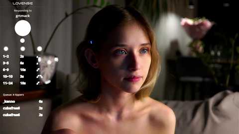 Media: A video of a topless woman with fair skin and shoulder-length blonde hair, standing indoors next to a dimly lit lamp. The background shows a blurred, dark room with a flower arrangement.