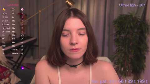 A video shows a young woman with shoulder-length brown hair, wearing a white camisole and a black choker, in a dimly lit room with a plant and a lamp in the background.