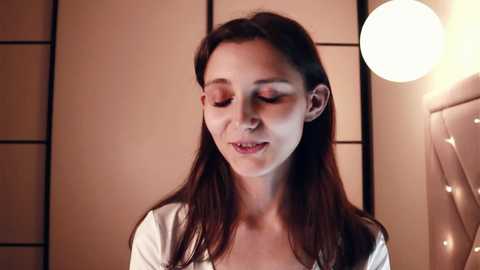 Video of a young woman with long dark hair, wearing a white top, smiling softly in a warmly lit, minimalist room with beige walls and a round pendant light.