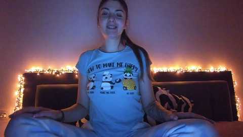 Media: Video of a smiling, light-skinned woman with long dark hair, sitting cross-legged on a bed with a pineapple-themed t-shirt, surrounded by warm, glowing fairy lights.