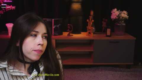 Media: Video of a young woman with long dark hair, wearing a striped shirt, sitting indoors with a dimly lit room in the background featuring a wooden console table, flowers, and a lamp.