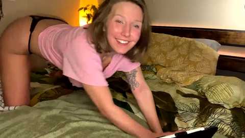 Media: A video of a smiling, light-skinned woman with shoulder-length brown hair, wearing a pink shirt and black panties, posing on a messy bed with green and brown bedding.