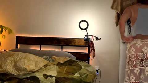 Media: Video of a woman in a sleeveless white dress adjusting a ring light on a wooden bed frame with a green quilt.