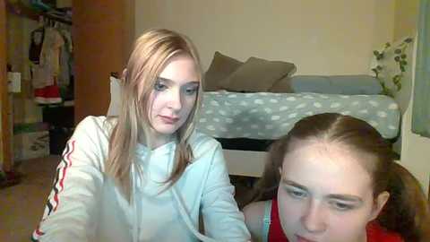 Media: Video of two young girls with light skin and straight hair, one in a light hoodie, the other in a red shirt, sitting in a bedroom with a bed, closet, and potted plant.