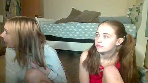 Media: A video of two young girls with long hair in pigtails, one in a red top and the other in a blue shirt, sitting on a carpeted floor in a bedroom with a white bed and polka-dot bedding.