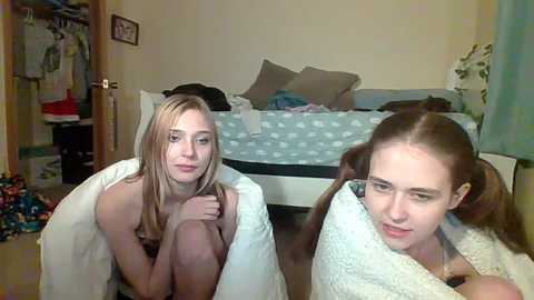 Media: Video of two young women, one blonde, one brunette, sitting on a bed, wrapped in towels, in a cluttered bedroom.