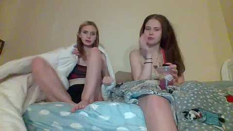 Media: Video of two young women, one blonde and one brunette, sitting on a bed, wearing casual clothing, drinking from a red cup, in a cozy, cluttered bedroom.