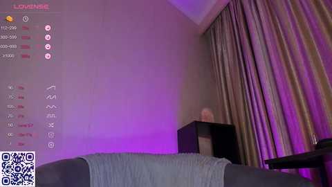 Media: A video of a dimly lit room with purple lighting. A person sits on a grey couch, wearing a grey sweater, with a large, dark TV stand behind them. The room has beige walls and heavy curtains.