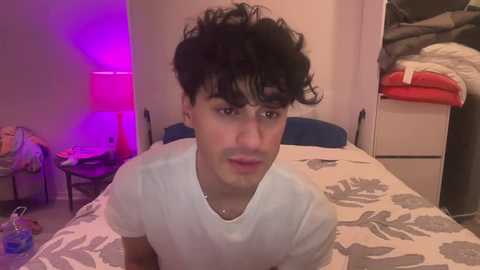 Media: Video of a young man with curly black hair, wearing a white T-shirt, sitting on a bed with floral sheets in a dimly lit, cluttered bedroom.