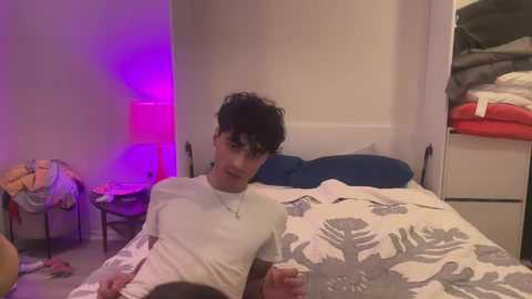 Media: Video of a young man with curly black hair, wearing a white T-shirt, lying on a bed with a gray and white floral-patterned duvet, in a dimly lit, cluttered bedroom with a purple light and stacked clothes in the background.