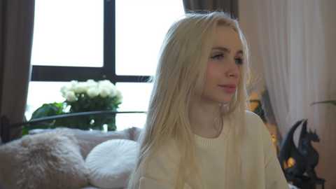 Media: Video of a young woman with long blonde hair, wearing a white sweater, gazing out a window with a floral arrangement in the background.