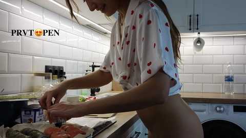 Media: Video of a slender, topless woman with long hair wearing a white, cherry-patterned shirt, preparing meat on a kitchen counter in a modern, white-tiled kitchen.