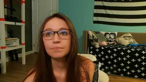 Video of a young Caucasian woman with straight brown hair and glasses, sitting in a teal-colored room with a flag, a cat, and a bed with a black and white patterned duvet.