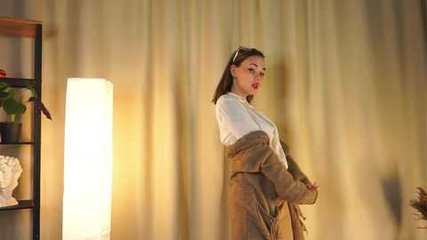 Media: Video of a young woman in a white blouse and brown jacket, standing in a softly lit room with a tall, cylindrical lamp, beige curtains, and a black shelving unit.