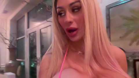 Media: Video of a blonde woman with long hair, wearing a pink tank top, looking surprised, in a modern room with glass doors and a framed painting.