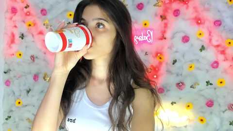 Media: Video of a young woman with long, wavy dark hair, wearing a white tank top, drinking from a white and red bottle. She stands against a floral-patterned wall with pink and yellow flowers.