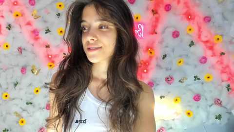 Media: Video of a young woman with long, wavy dark hair, wearing a white tank top, standing against a vibrant floral backdrop with pink, yellow, and red flowers.