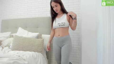 Media: Video of a young woman with light skin and straight dark hair, wearing a white crop top and tight gray leggings. She stands in a modern bedroom with white brick walls and a light-colored bed.