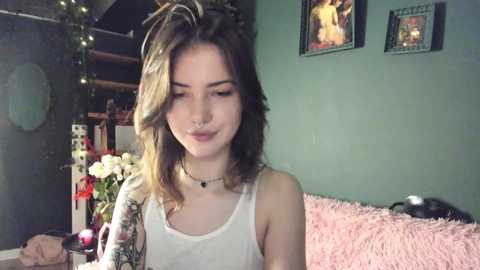 Media: Video of a young Caucasian woman with shoulder-length brown hair, wearing a white tank top and black choker, standing in a dimly lit room with pastel green walls and a pink shag rug.