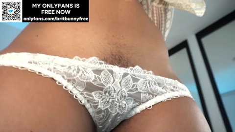 A close-up video of a woman's lower abdomen, showing a white lace thong with floral patterns. The background includes a mirror and a partially visible window, with text promoting an OnlyFans account.