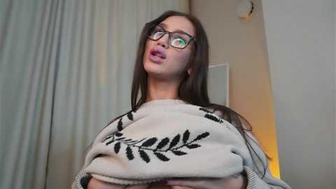 Media: Video of a young woman with fair skin, long brown hair, and glasses, wearing a cream sweater with a black leaf design. She has a surprised expression. Background shows a white wall and a partially visible curtain.