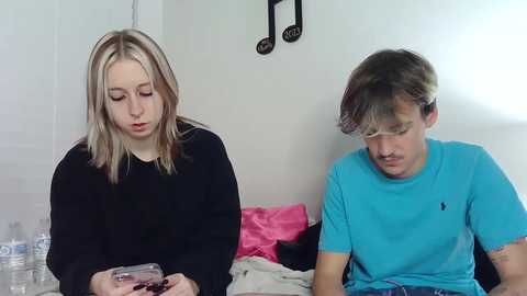 Media: Video of a young blonde woman in black, holding a phone, next to a young man in a blue shirt, both seated on a bed with pink sheets and a water bottle in the background.