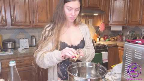 Media: Video of a young Caucasian woman with long brown hair, wearing a black lace bra and beige cardigan, stirring a metal pot on a stove in a cozy kitchen with wooden cabinets.