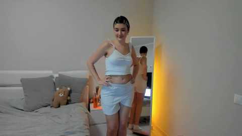 Media: Video of a young woman with fair skin and dark hair in a ponytail, wearing a white crop top and shorts, standing in a minimalist bedroom with a bed, teddy bear, and a mirror.