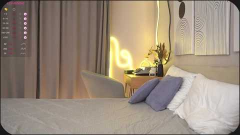 Video of a modern bedroom with light gray bedding, white pillows, and a bedside table holding a lamp and vase. Minimalist decor, wall art with circular patterns, and soft lighting.