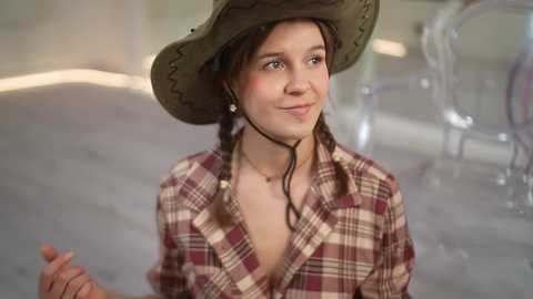 Media: Video of a young woman with light skin, wearing a green cowboy hat, plaid shirt, and pearl earrings, smiling, in an indoor setting with transparent chairs and a blurred background.